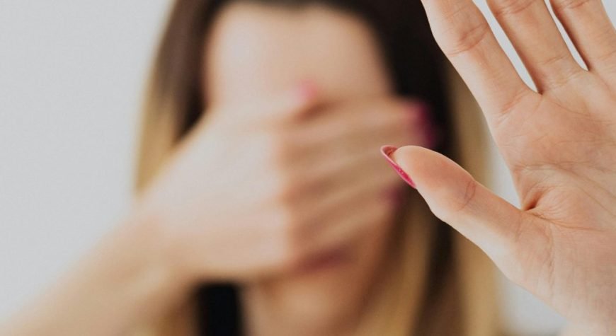 A woman in a gray shirt covers her face with her hand in a stop gesture, evoking a sense of fear and protection.