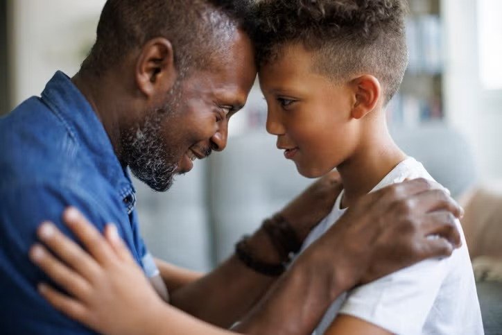 Black father sitting quietly on the porch steps at dusk while his child plays nearby, representing fathers who carry stress in silence